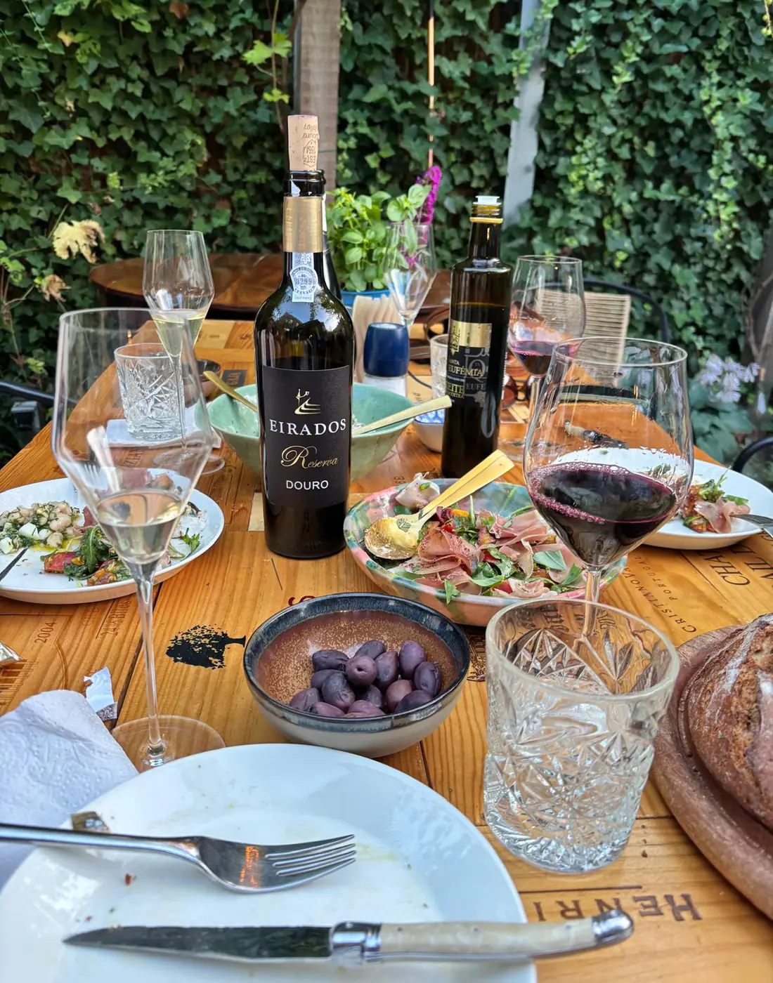 A garden table set with an open bottle of Eirados Reserva, presunto, olives, bread and glasses of red and white wine.